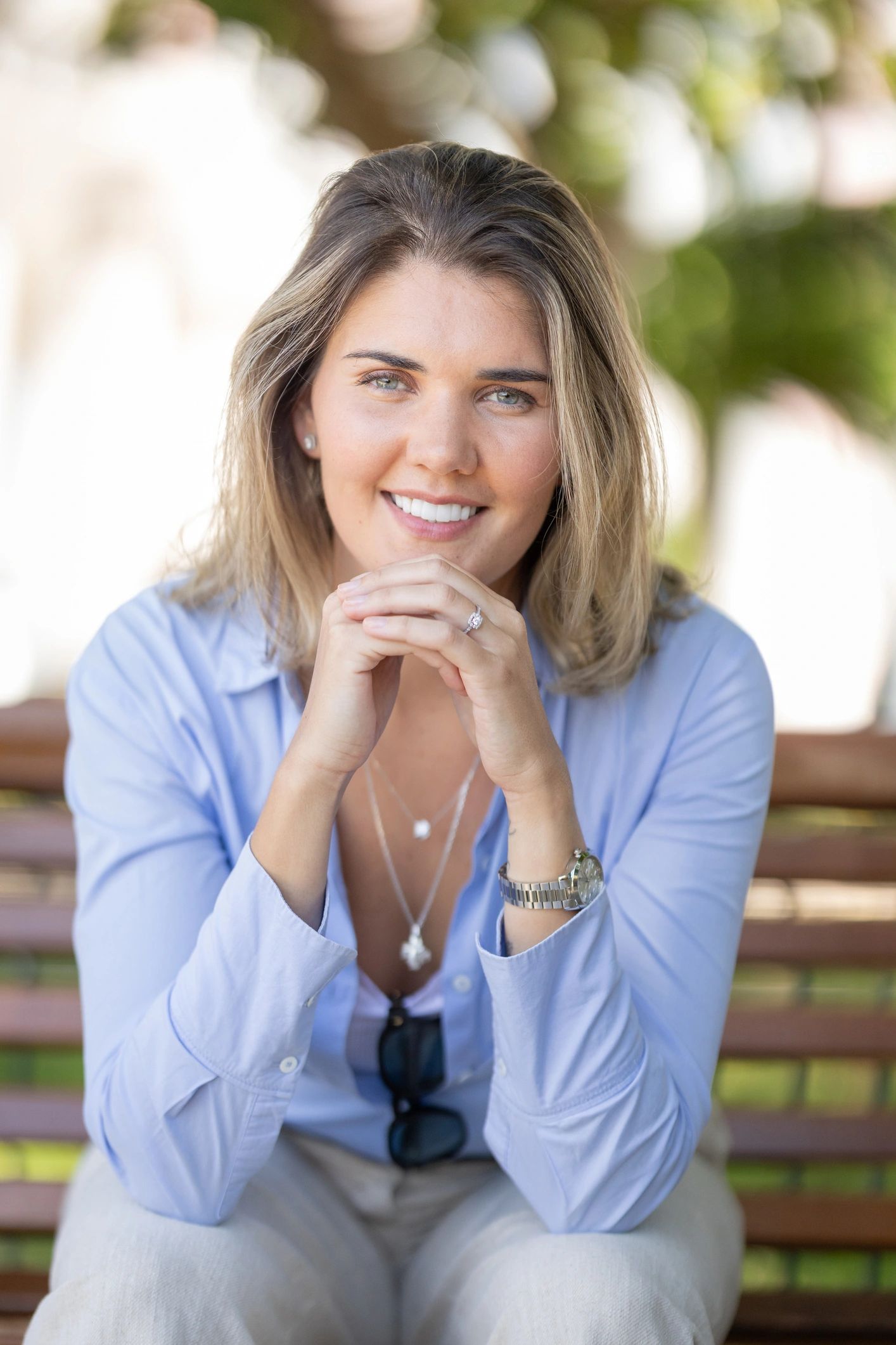 Woman sits on a bench in a park with a smile and hands clasped in front of her while wearing a light blue shirt and jewelry